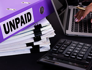 Purple binder labeled UNPAID on stacked papers beside a calculator and laptop.